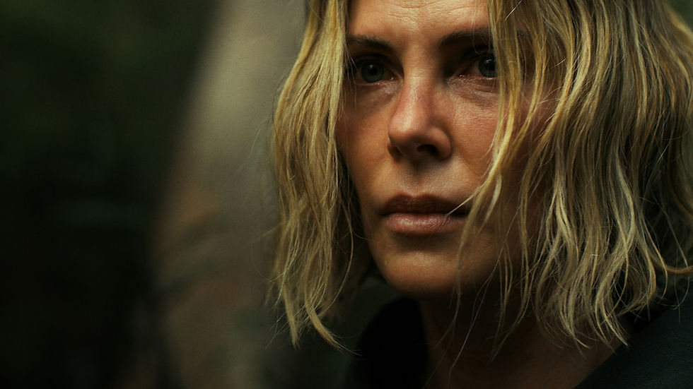 Close-up of a person with short, blond hair in a forest, looking serious. The background is blurred with earthy tones.