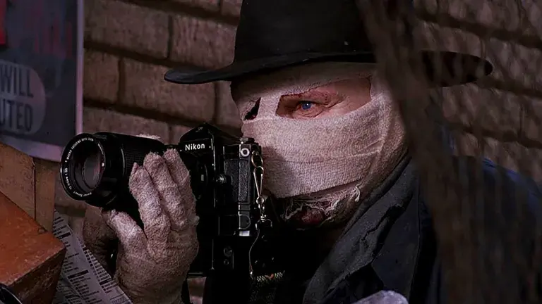 Bandaged man in a black hat peers through a Nikon camera in a brick alley, creating a tense, eerie mood.
