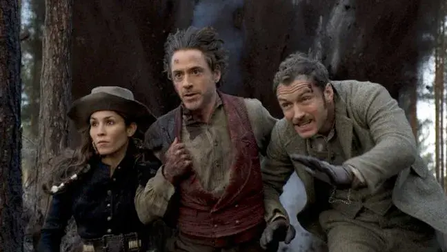 Three anxious people in period clothes crouch and run through a dark forest, looking offscreen.