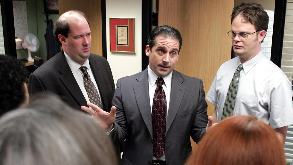 Three office workers in suits listen as a man gestures mid-speech in a cluttered office, tense and serious.