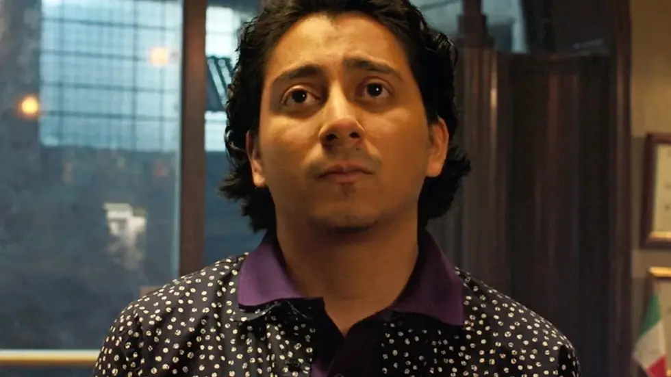 A person with brown hair looks contemplative in a room with framed certificates. They wear a dark shirt with white dots. Soft indoor lighting.