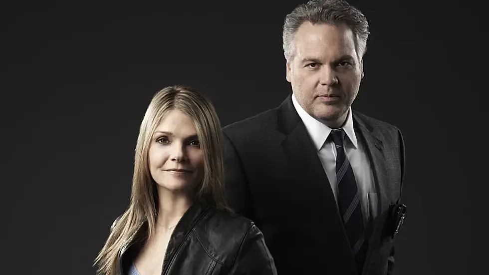 Serious man in suit stands behind a woman in a black jacket against a dark studio background.