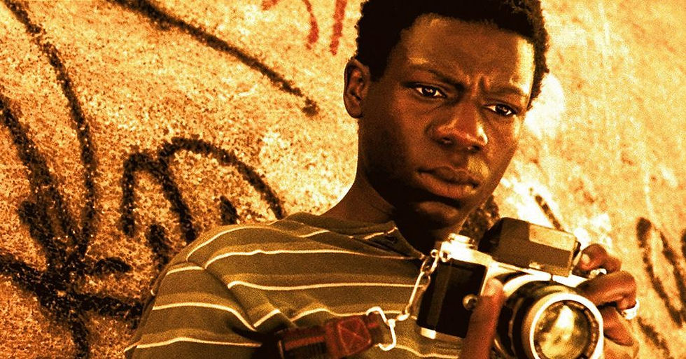Young man with a camera stands before a graffiti wall, looking serious in warm orange light.