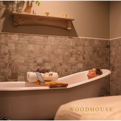 Woman relaxing in a white bathtub with a laptop and drink in a tiled spa; candles on a shelf.