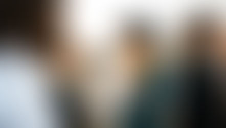 People on a rooftop, smiling and talking while holding wine glasses. Tattoos and a black hat visible. Cityscape in background, sunset light.