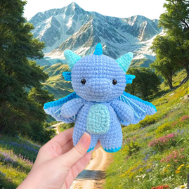 A light blue crochet dragon with teal horns, blue spikes on its back, blue gills below the horns, a teal belly, and light blue wings tipped in blue and accented with teal lines.