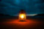 Lit lantern on a sandy beach at dusk, emitting a warm glow against a dark, cloudy sky, creating a calm and serene atmosphere.