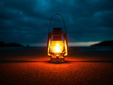 Lit lantern on a sandy beach at dusk, emitting a warm glow against a dark, cloudy sky, creating a calm and serene atmosphere.