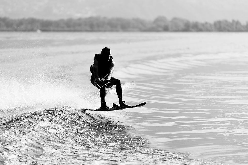 Black-and-white photo of a wakeboarder carving across a calm lake, spraying water with a blurred shoreline behind.