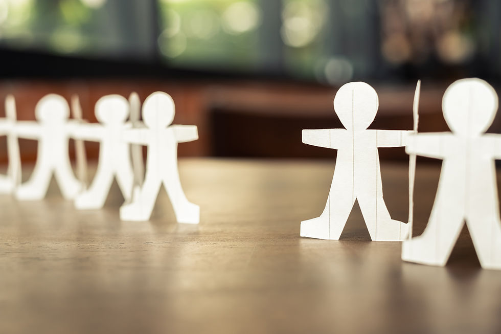 Paper chain figures on a wooden table, with a blurred green and brown background. The figures are disconnected and reaching for each other
