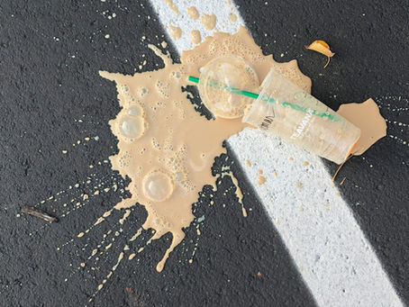 Spilled coffee on asphalt next to a white line. Transparent cup with a green straw lies on the ground, creating a messy scene.