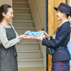 Woman receives a package of Yakult yogurt from a smiling delivery woman in uniform at her doorstep. Stairs in the background.