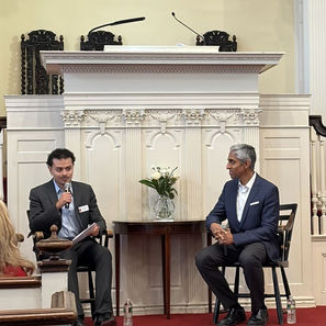 Two men in suits in front of a church pulpit have a conversation, one speaking into a microphone beside a flower vase.