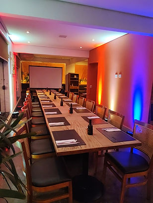 Uma mesa para um jantar corporativo. Ao final da mesa há um telão para fazer as projeções da empresa e no chão canhões de luz para tornar o ambiente agradável.
