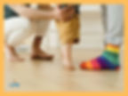 Bambino che muove i primi passi sostenuto da due adulti, uno con calzini arcobaleno. Immagine simbolica del passaggio e del cambiamento nel percorso dell’allattamento. Life Attività e servizi psicologici