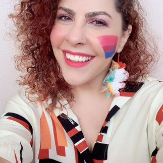Foto da autora Brenda Zulp em estilo selfie. Mulher banca com cabelos cacheados vermelhos jogados para o lado esquerdo. Ela usa batom cor de rosa, está sorrindo e veste uma camisa clara de abotoar com estampa geométrica nas cores vermelho, preto e laranjado e usa um grande brinco na forma de um unicórnio de pelúcia. Seu rosto está virado para o lado esquerdo e sua bochecha direita está pintada com as cores da bandeira bissexual: rosa, roxo e azul. O fundo é branco.