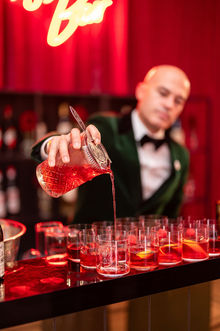 a smartly dressed barman in a tuxedo and bow tie strains a cocktail into a tumbler on a black bar with a neon light behind at a corporate brand event with multiple drinks ready for a slick service