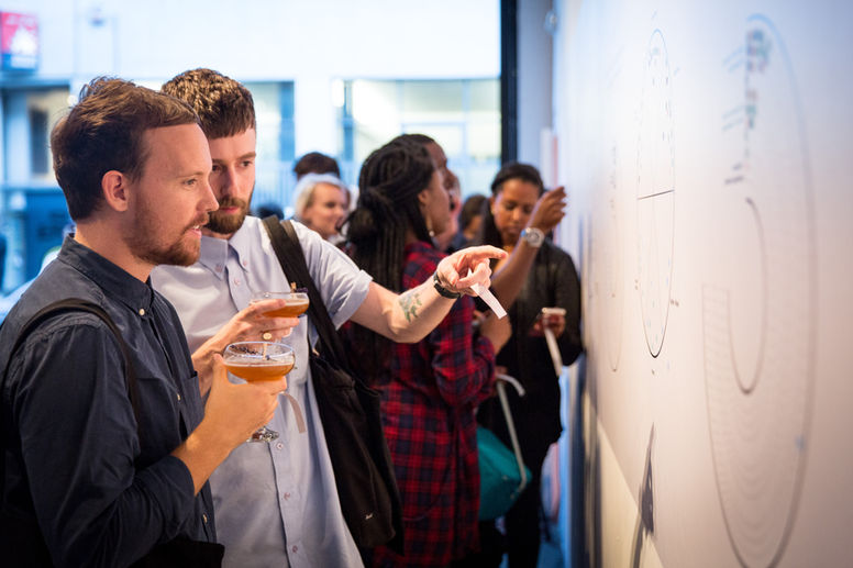 Two young men holding martini glasses with a cocktail in discuss an art piece on the wall in a light art gallery in east London. The two men look as though they have recently finished work in a media or design role.