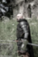 Matt Whitfield In black medieval armour, looking across a battelfield. he has a sword sheathed in it