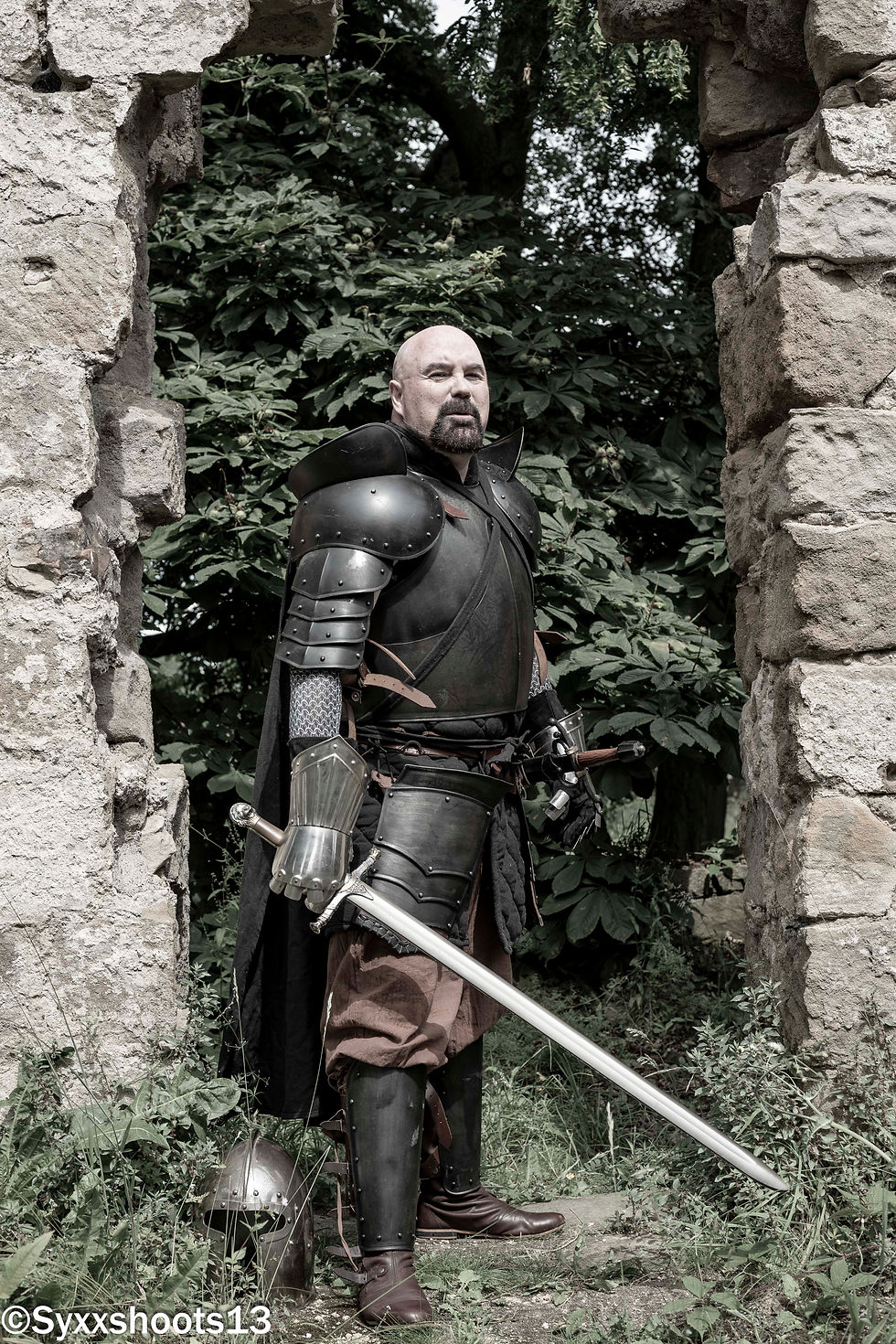 Matt Whitfield In black medieval armour, outside a medieval building. He has his sword drawn..