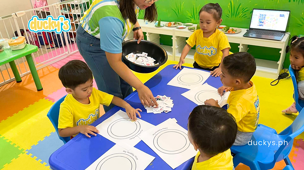 Teacher and preschoolers in yellow Ducky's shirts sort food cards on healthy plate worksheets in a colorful classroom.