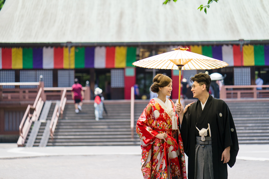 赤い色打掛姿の花嫁と黒紋付袴の新郎が、喜多院の前で和傘を差しながら見つめ合う様子
