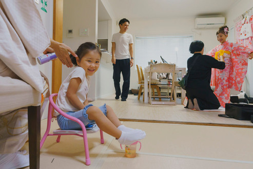 七五三の準備シーンで、小さな女の子が椅子に座って笑顔で髪を整えてもらい、奥では大人の女性がピンクの着物に着付けを受けている。家族が見守る中でのお支度の様子。