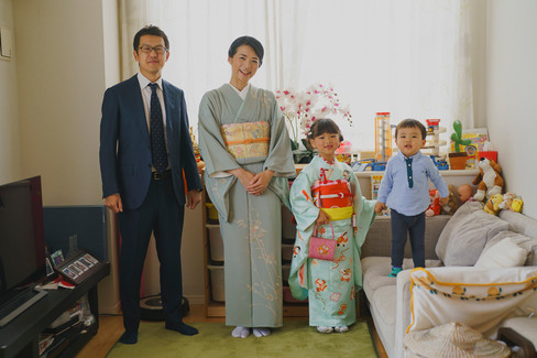 七五三撮影当日の朝、自宅のリビングで着物姿の母と娘、スーツ姿の父、普段着の弟が並び、記念写真を撮っている。おもちゃや花が彩る家庭的な空間。
