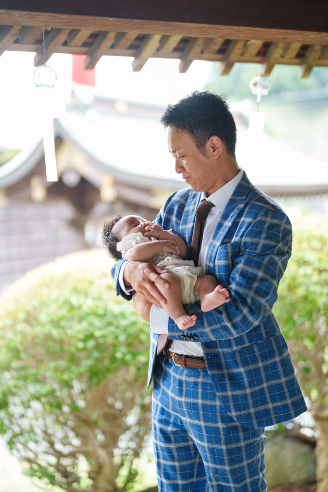 神社の境内で、赤ちゃんを優しく抱く父親の姿。風鈴が揺れる中、穏やかに見つめ合う父子のひととき。