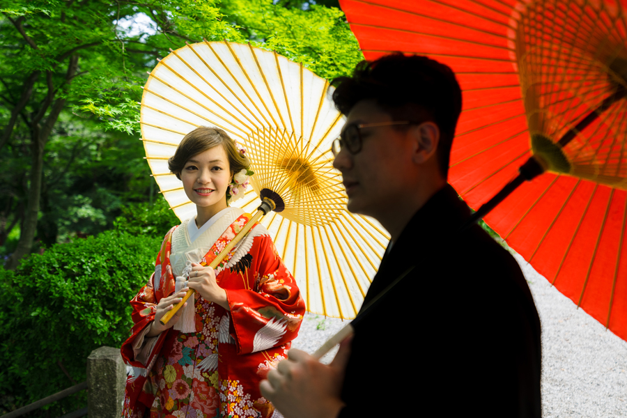緑に囲まれた庭園で、和傘を持つ花嫁が微笑み、新郎が見守るシーン