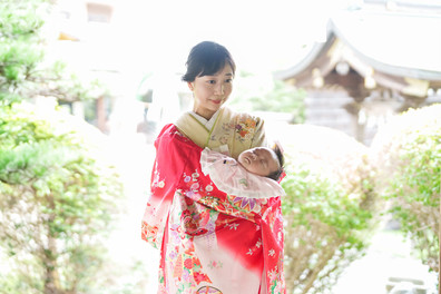 柔らかな光の中、赤い祝い着をまとった赤ちゃんを抱く母親が神社境内にたたずむ、優しい雰囲気のポートレート。