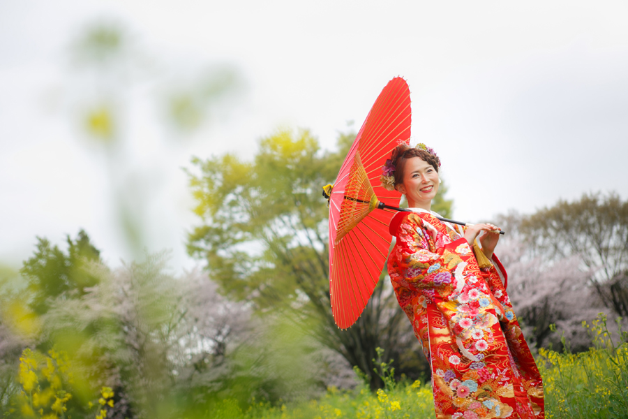 赤い和傘を手に、満開の桜と菜の花に囲まれて微笑む花嫁のソロショット