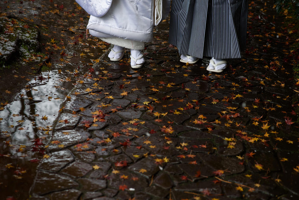 雨上がりの石畳に落ち葉が散る中、白無垢姿の足元を写した印象的な和装写真