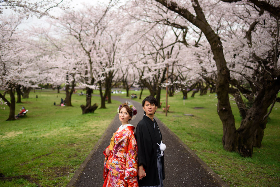 桜並木を背景に立ち、少しクールな表情で見つめる和装姿の新郎新婦