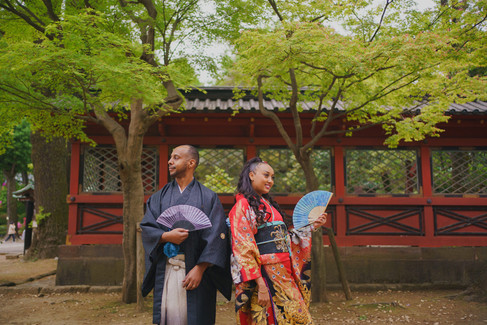 Tokyo wedding photographer capturing couple in kimono holding fans in front of red wooden wall at Nezu Shrine Japan