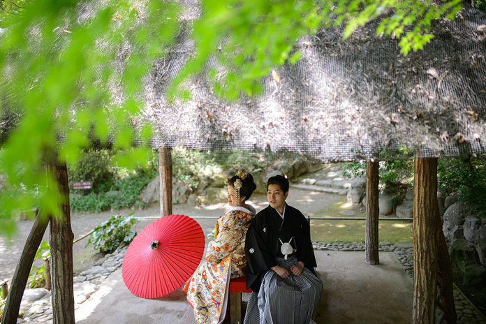 茅葺き屋根の東屋で寄り添う和装の新郎新婦と赤い和傘｜伝統的な前撮り写真