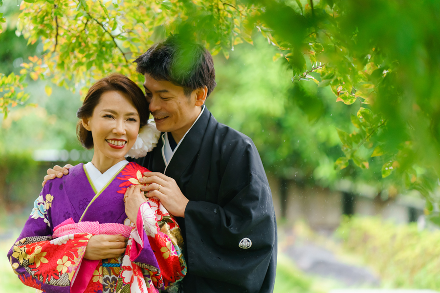 緑に囲まれた芝公園で、和装の新郎新婦が手を取り合い笑顔を見せる様子