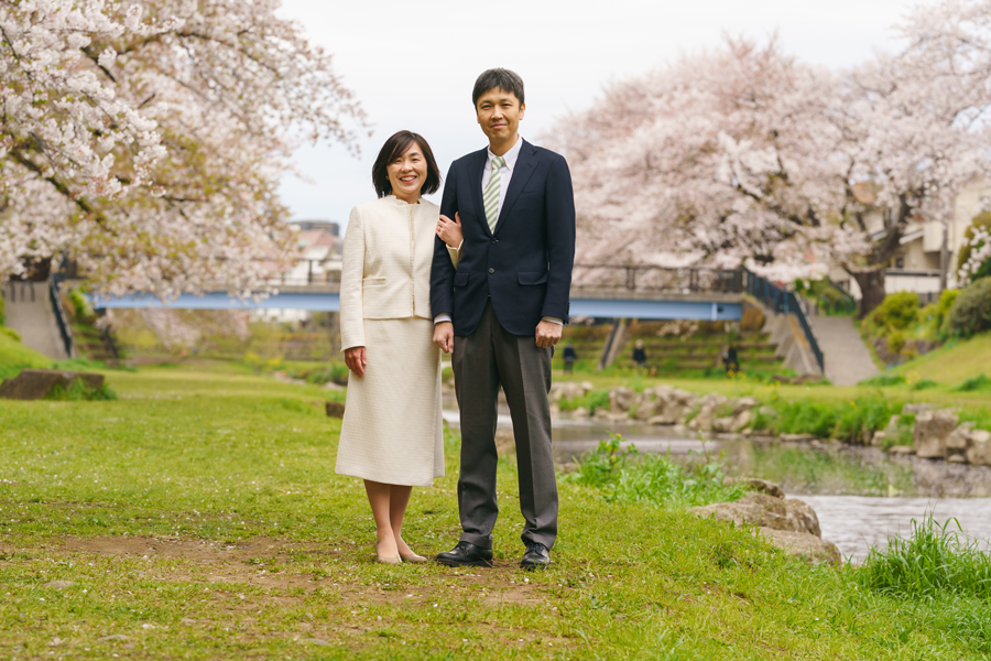桜並木と川を背景に、腕を組んで立つ夫婦の春のロケーションフォト