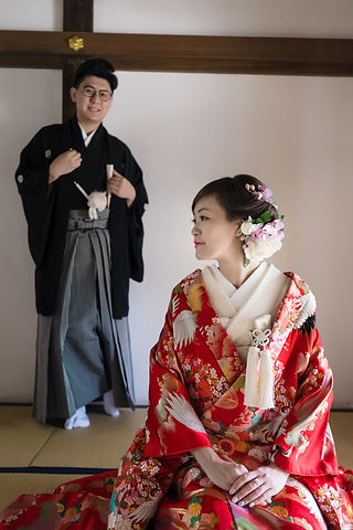 赤い色打掛姿の花嫁が前に座り、黒紋付袴の新郎が後ろから見守るポートレート（川越・和室での撮影）