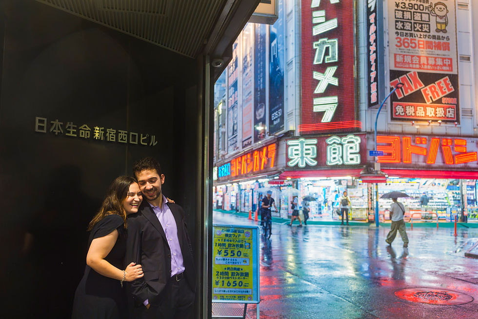 Laughing honeymoon couple beside neon-lit electronics stores on a rainy night in Shinjuku