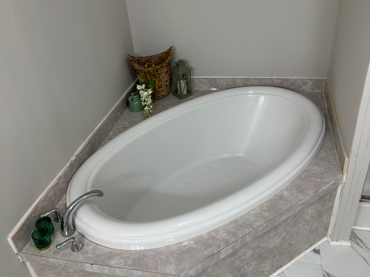 Oval refinished bathtub in a marble-tiled bathroom with green candles, wicker basket, green lantern, and white flowers. Calm and inviting space.