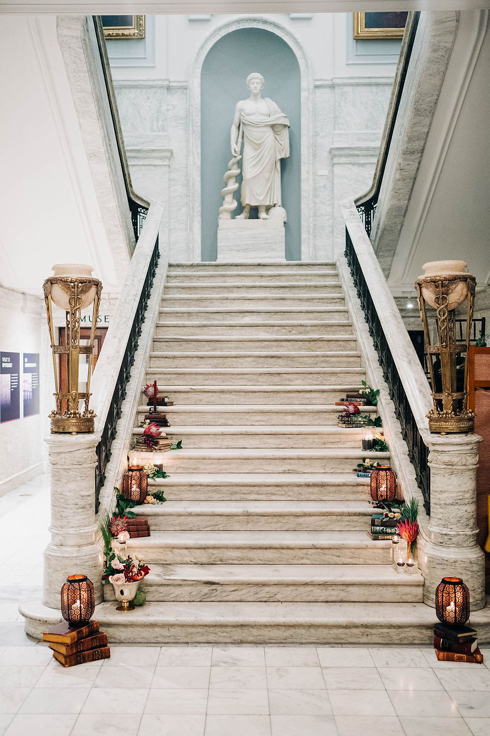 Elegant marble staircase with a statue at the top. Books, flowers, and lamps line the steps, creating a warm, inviting atmosphere.