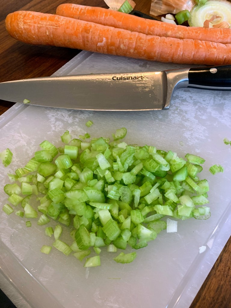 Chopped celery on a white cutting board with a Cuisinart knife and whole carrots nearby. Wooden table background.