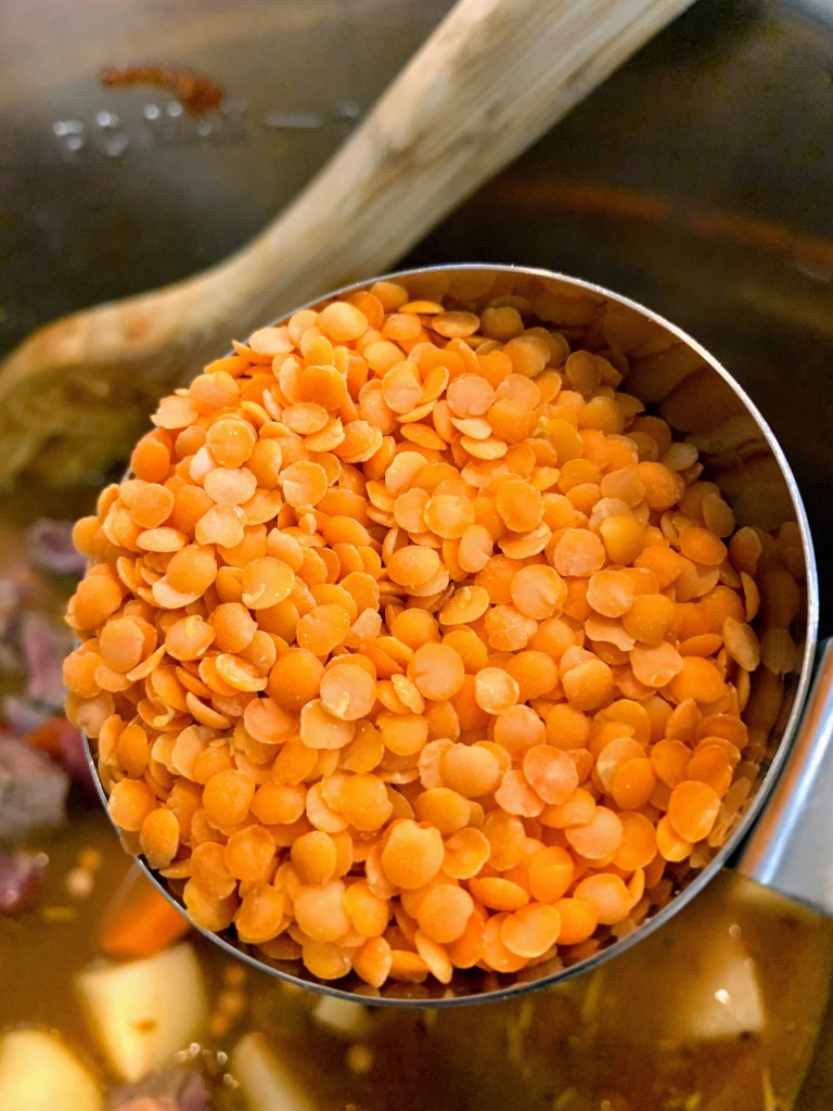 A cup of bright red lentils held over a pot with a wooden spoon. Background includes chopped vegetables submerged in broth.