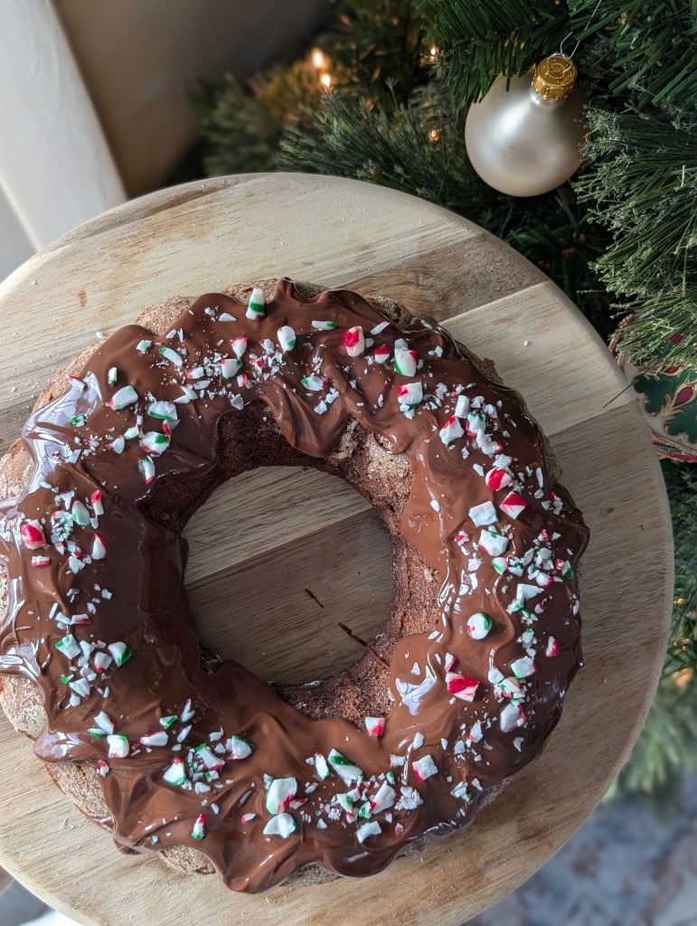 Chocolate-covered cake with crushed peppermint on a wooden board. Background features a Christmas tree with a gold ornament, creating a festive mood.