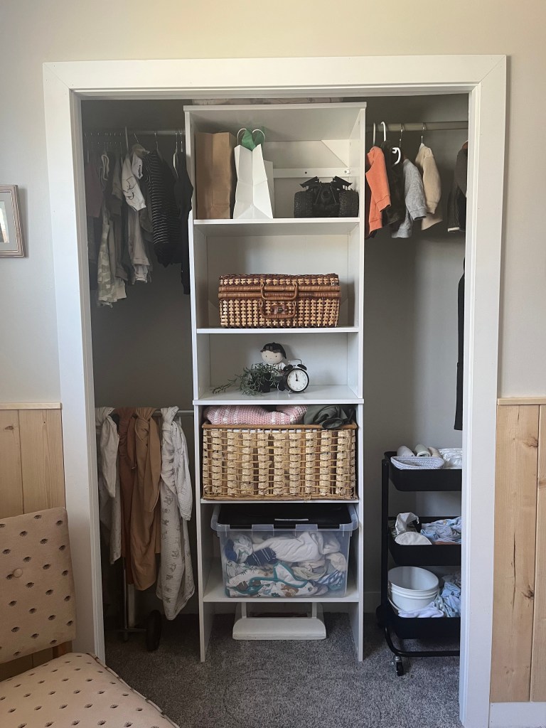 Organized closet with clothes, wicker baskets, a clock, and storage bins. Neutral tones, cart with bowls. Cozy, tidy setting.