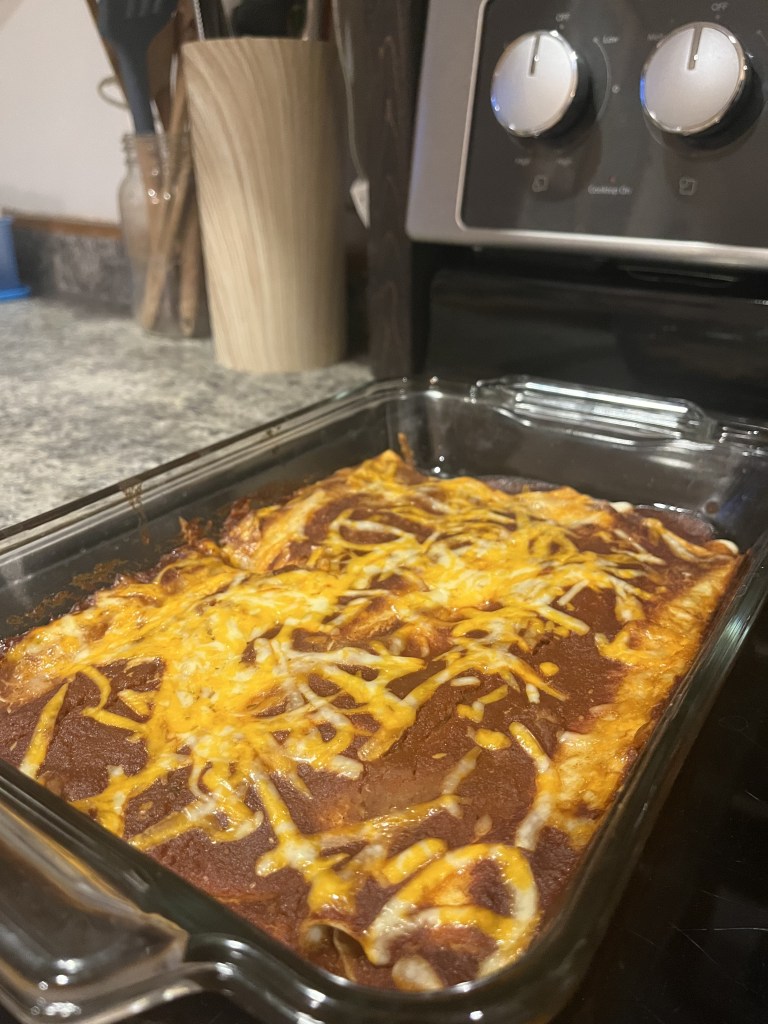 Baked enchiladas with melted cheese in a glass dish on a kitchen countertop. Stove knobs visible in the background. Cozy kitchen setting.