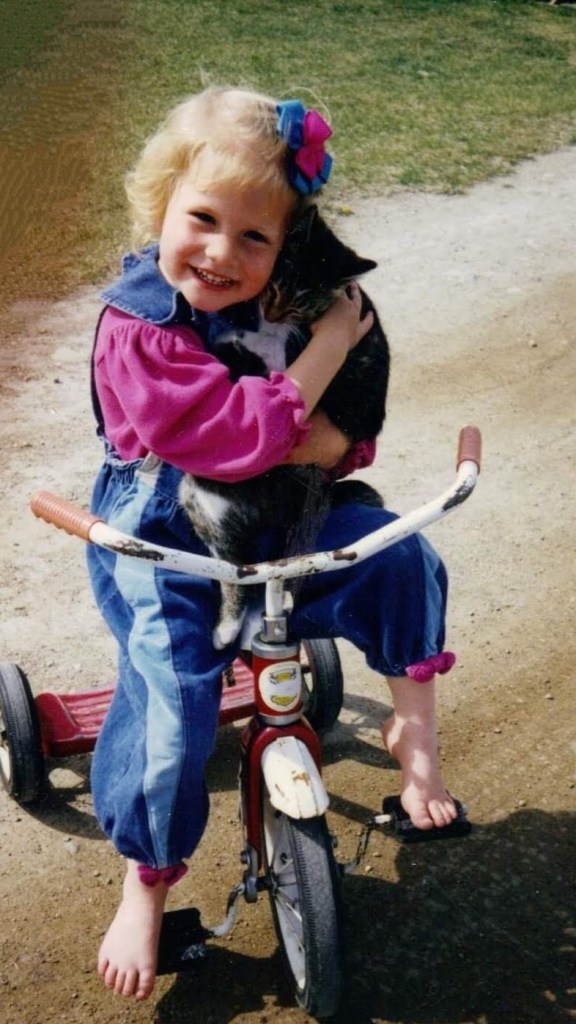 Young child with blonde hair and a bright bow hugs a cat on a tricycle. Wearing a pink and blue outfit, they smile outdoors on a sunny day.