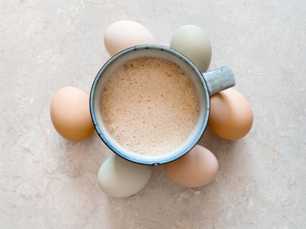 A blue cup of frothy coffee on a beige surface, surrounded by six pastel-colored eggs in a circle. Calm and cozy mood.