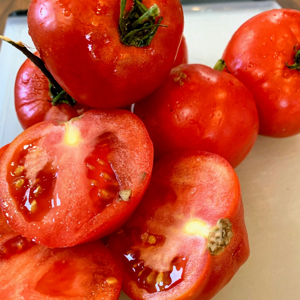 Fresh red tomatoes on a white surface, some whole and one sliced open, showing juicy seeds. Bright and vibrant colors.